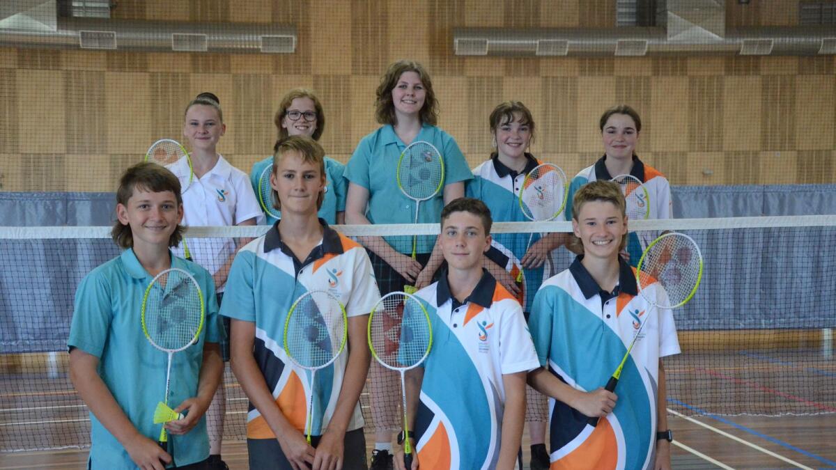 School students stand in front and behind a badminton net, smiling and holding racquets. 